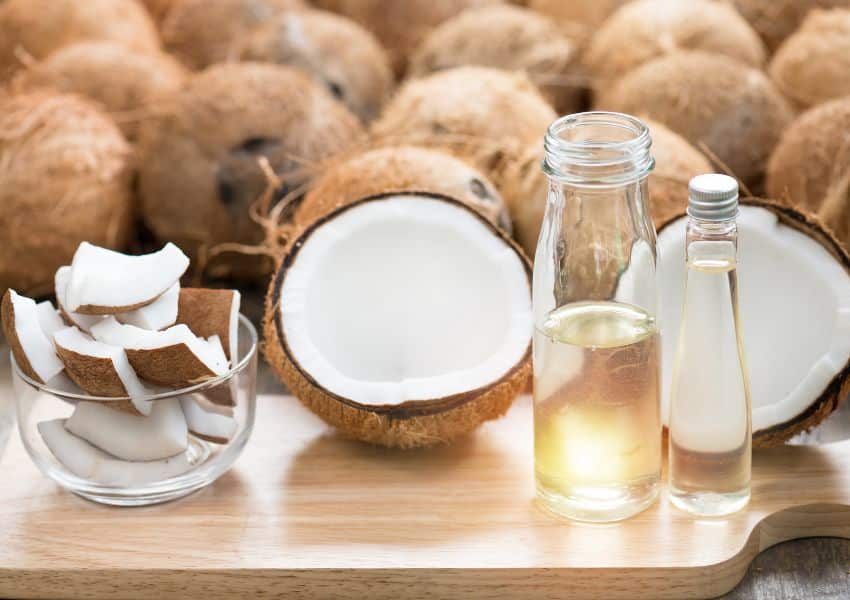 Um coco partido ao meio, agua de coco em uma garrafa, lascas de coco em um copo ao fundo vários cocos descascados