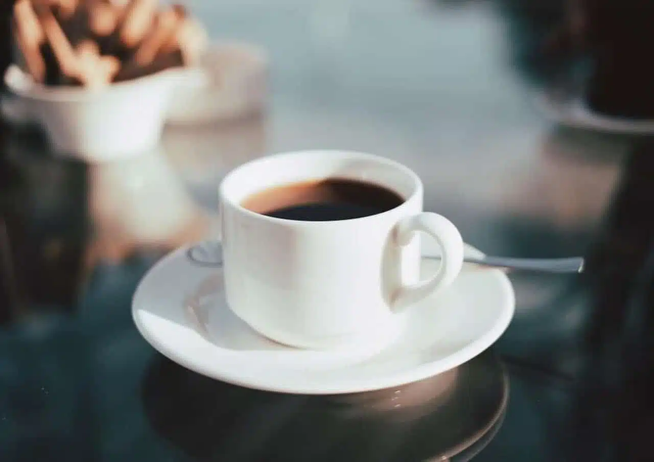 Copo alto com iced latte sobre gelo, ao fundo uma cafeteira e grãos de café espalhados na mesa