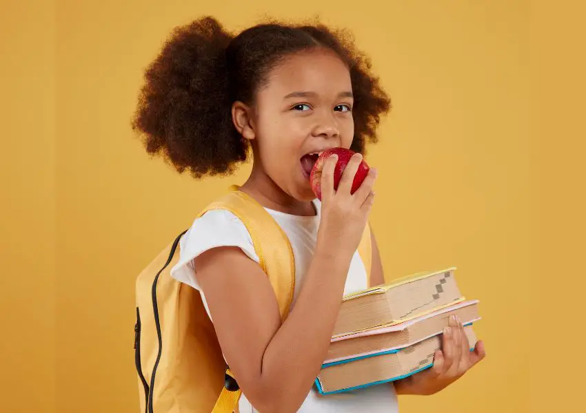 Crianca menina comendo maca e segurando livros escolares