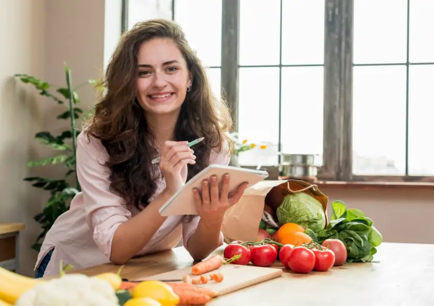 Mulher sorrindo com papel e caneta na mao muitas frutas e verduras